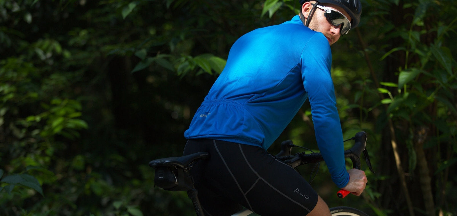 Cyclist wearing a blue long-sleeve jersey and black cycling shorts, riding outdoors and looking back on a forest road